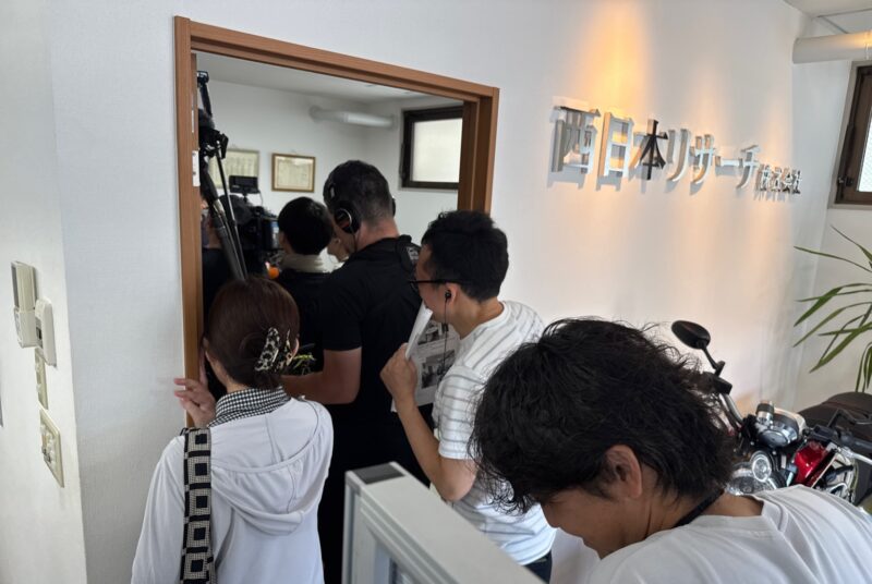撮影中の西日本リサーチ株式会社のオフィス風景。番組スタッフが集まり、機材と共に撮影準備を進めている様子。